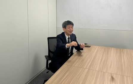 Smiling man in a dark suit sits at a wooden conference table in a small office, gesturing with his hands.