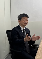 Man in a dark suit seated at a desk, gesturing with both hands as if speaking in an office setting.