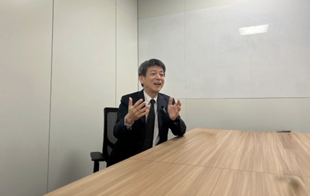 Man in a suit sits at a conference table in a modern office, gesturing as he speaks during a meeting or interview context.