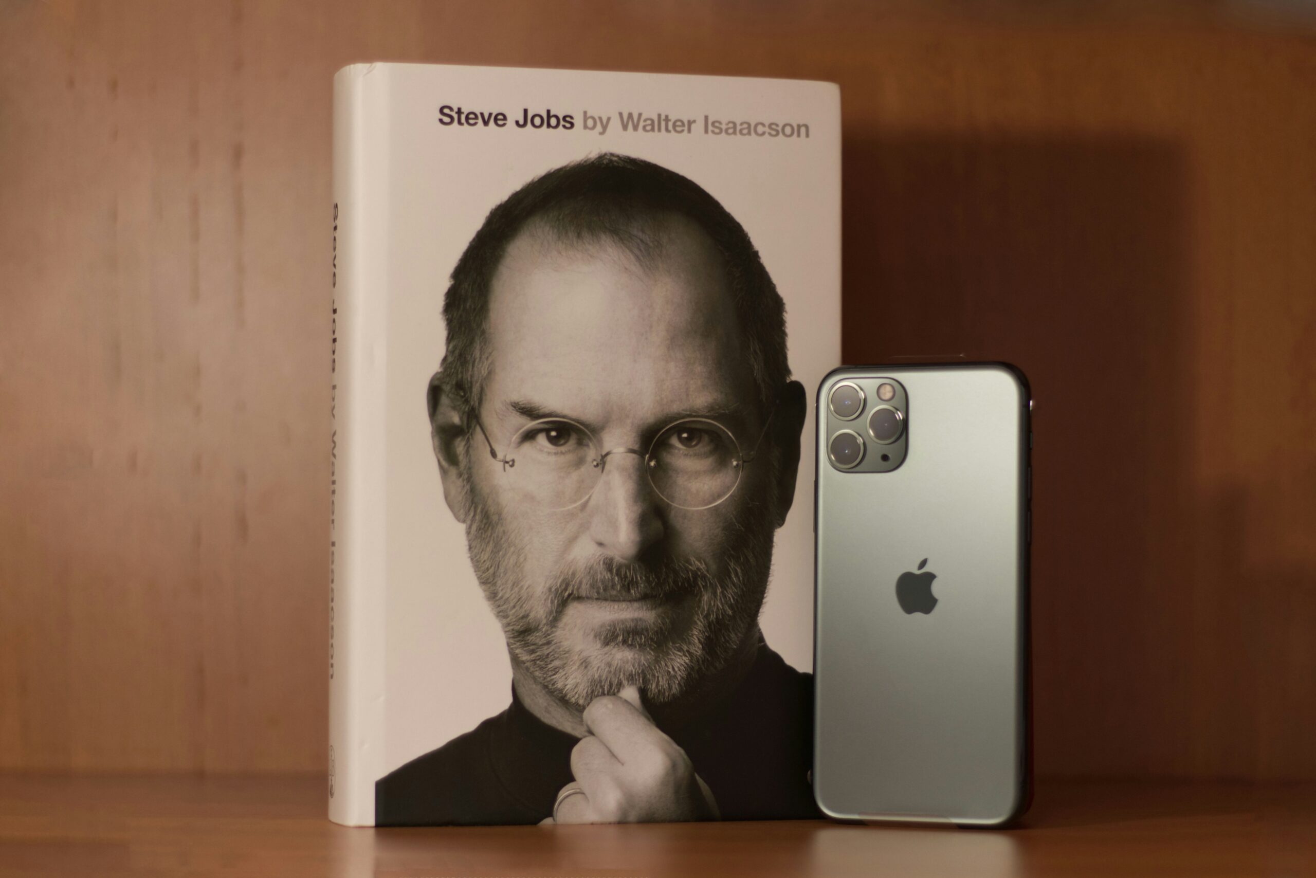 Biography cover'Steve Jobs by Walter Isaacson' next to a silver iPhone on a wooden shelf.