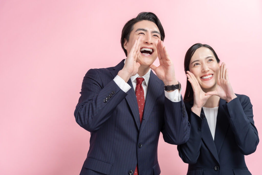 呼びかけるビジネスマン男女 - SEMICON.TODAY Two coworkers in navy suits cheerfully cupping hands around their mouths against a pink background, celebrating together.
