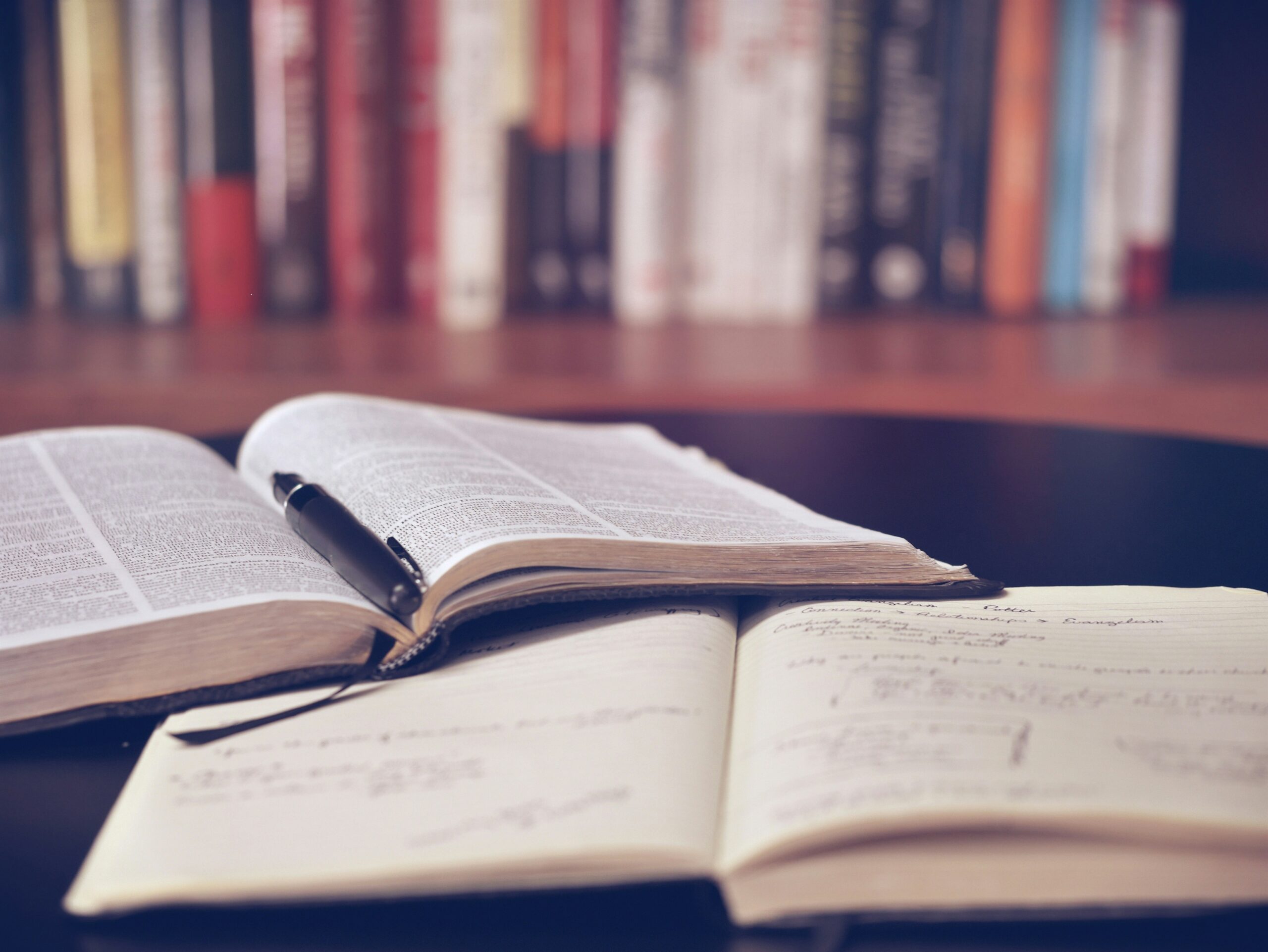 Open book with a pen resting on its pages on a desk, blurred bookshelf in the background.