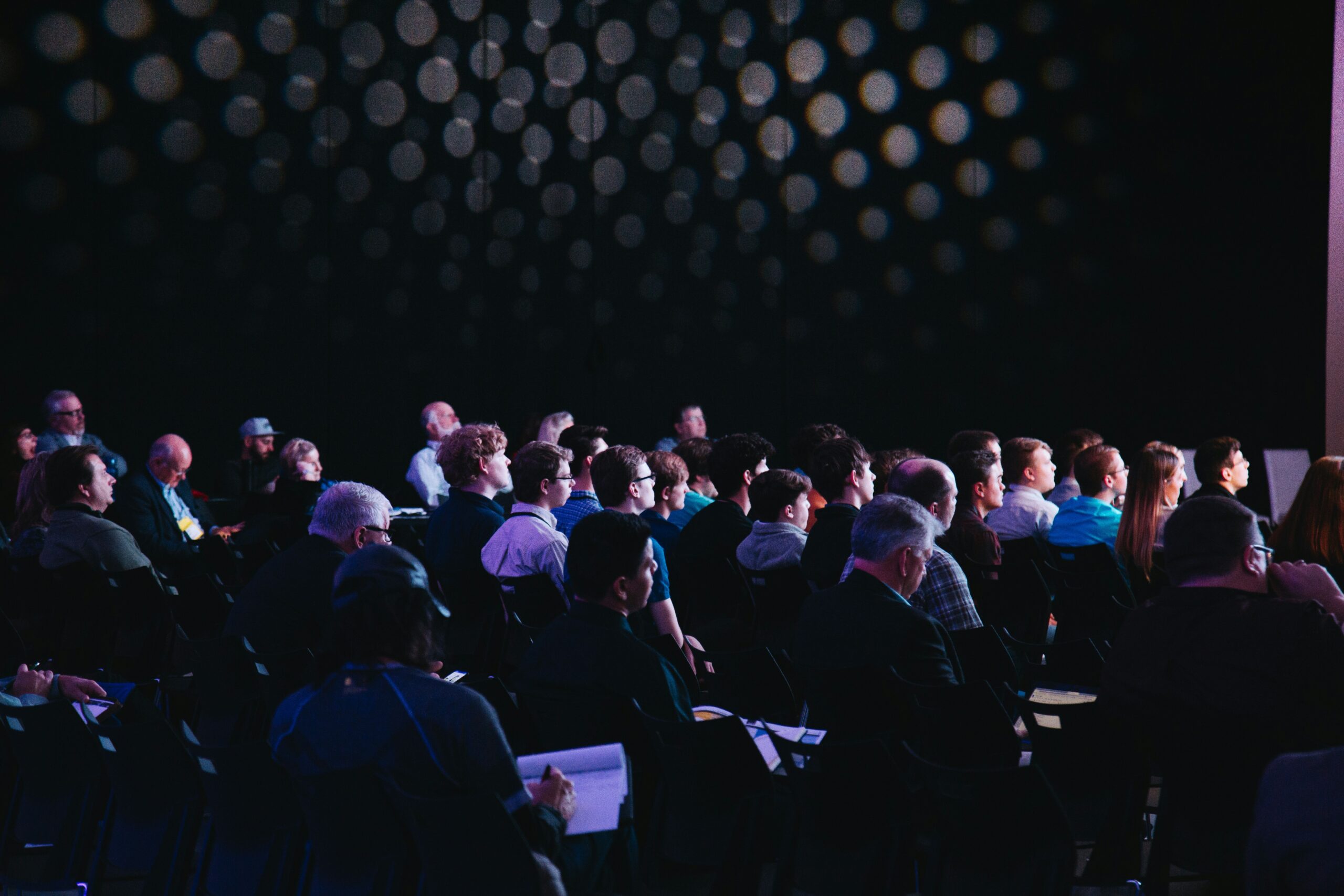 headway-F2KRf_QfCqw-unsplash - SEMICON.TODAY Audience seated in a dim theater watching a presentation, with blurred circular light bokeh overhead. (informative)