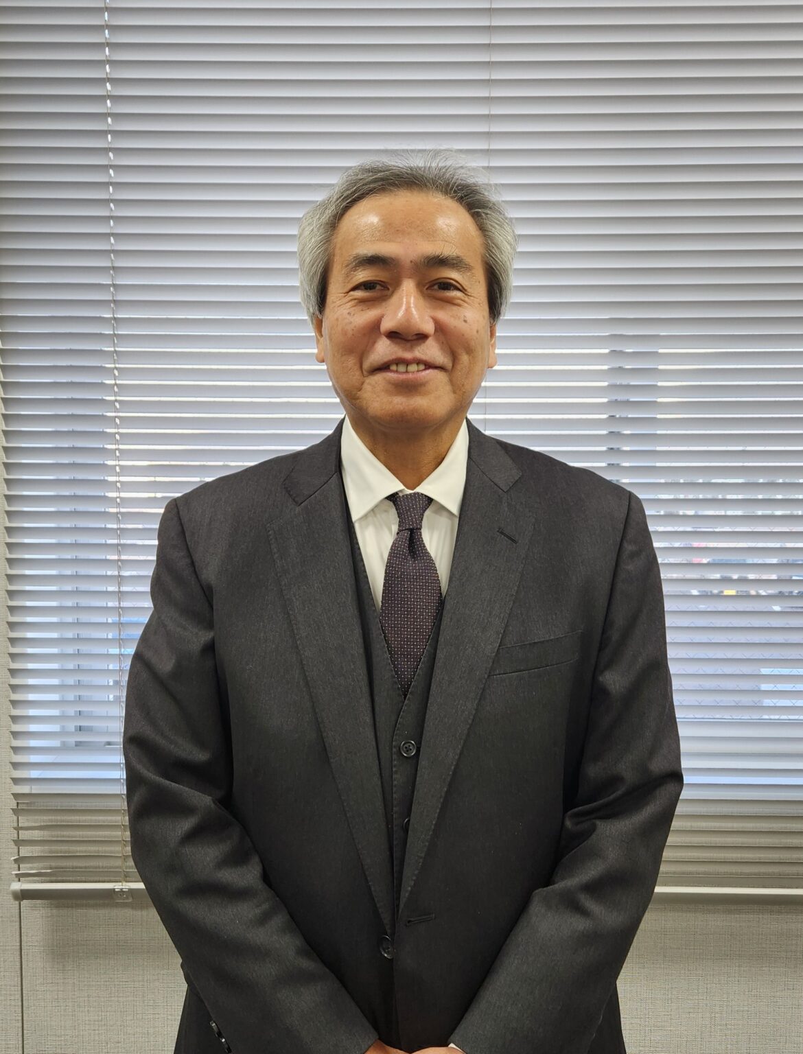 Older man in a dark suit and purple tie standing in front of office blinds, smiling.