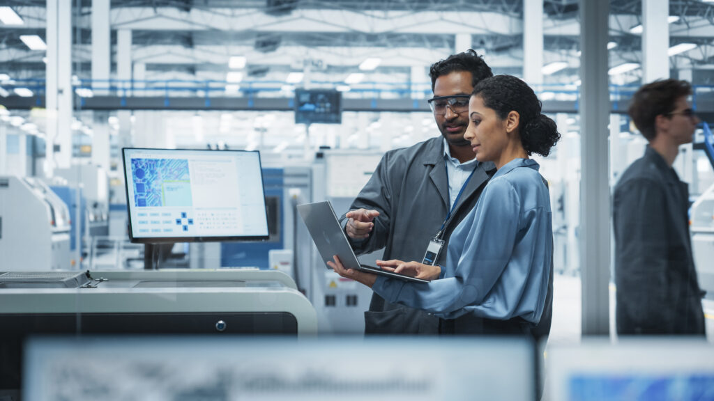 Indian Male Industrial Engineer And Hispanic Female Supervisor Using Laptop And Talking At An Electronics Factory. Man Using Soldering Jet Printer, Explaining Process Behind Production Of Motherboards - Semicon.TODAY