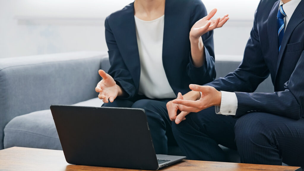 ロビーでノートパソコンを見ながら会話する男女ビジネスパーソンのグループ - Semicon.TODAY