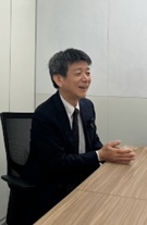 Man in a dark suit seated at a wooden conference table, speaking with hands resting on the table in a meeting room.”,