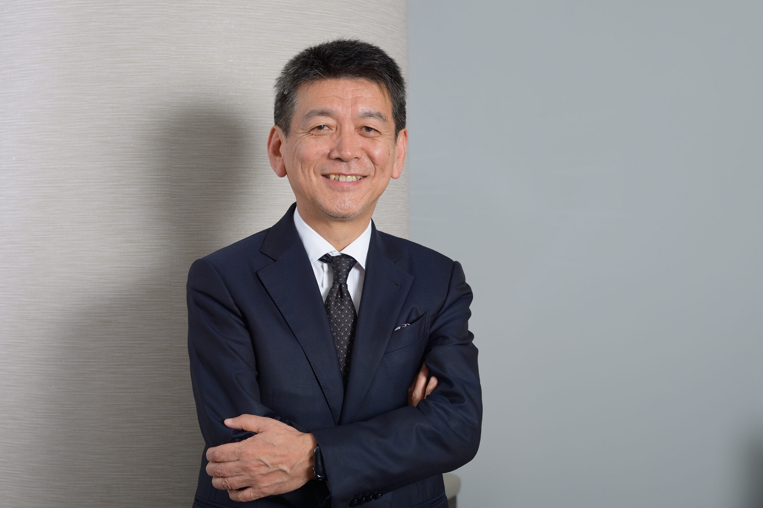 Smiling man in a dark suit with a patterned tie, arms crossed against a textured wall.