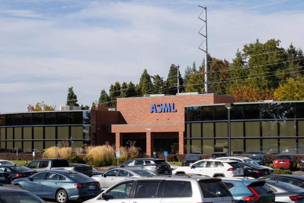 ASML office building with brick exterior and prominent blue ASML sign, surrounded by a parking lot and trees under a blue sky with power lines overhead