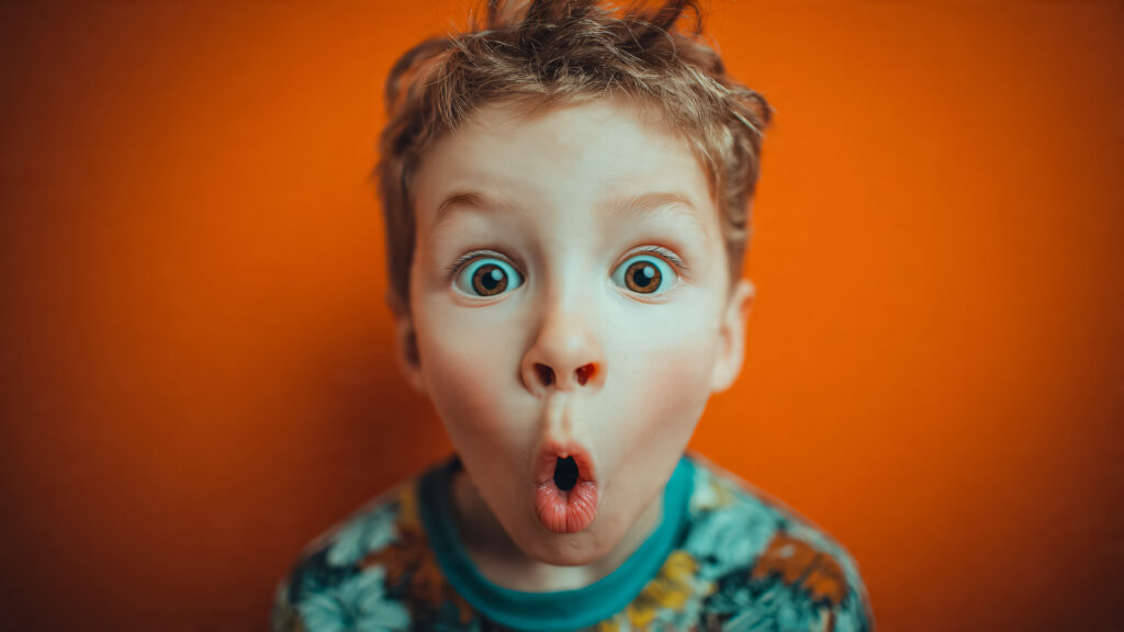 Petit garçon très surpris avec la bouche ouverte et des grands yeux - SEMICON.TODAY Close-up of a young boy with wide blue eyes and puckered lips against an orange wall, looking surprised.