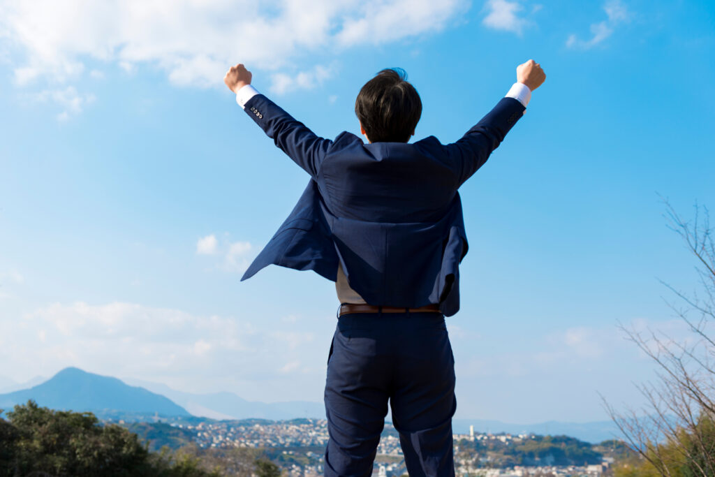 Person in a dark blue suit stands with arms raised in a victory pose, back to the camera, overlooking a valley and mountains under a blue sky.