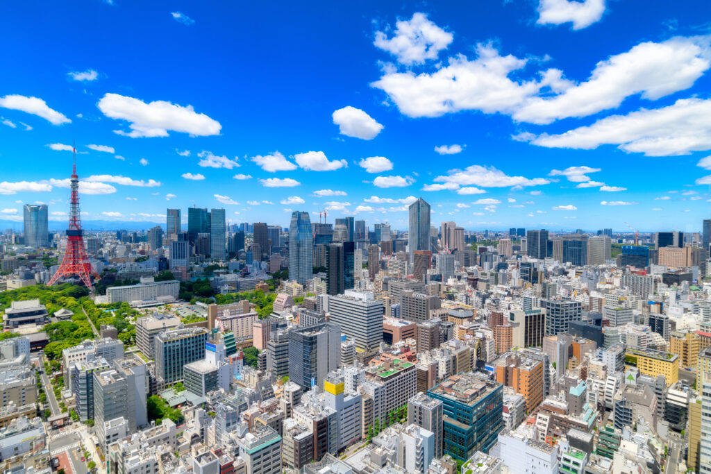 東京の都市風景 - SEMICON.TODAY Wide daytime cityscape with numerous skyscrapers and a red-and-white Tokyo Tower on the left, under a bright blue sky with scattered white clouds.