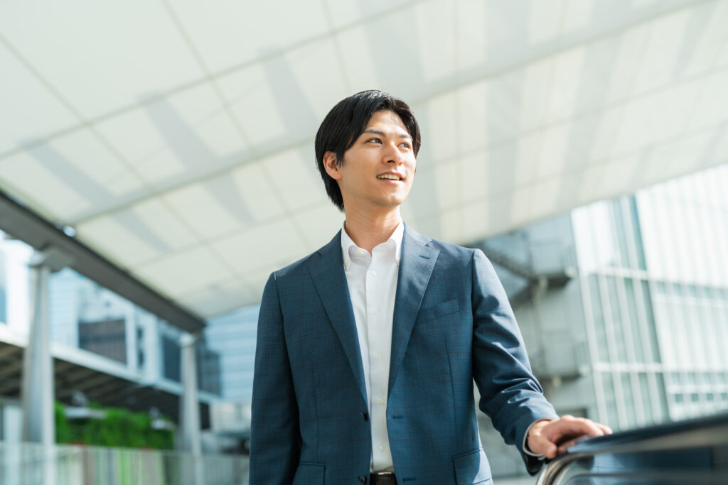 Young Businessman - SEMICON.TODAY Professional man in a blue suit standing on an outdoor escalator near a glass building, looking to the right.