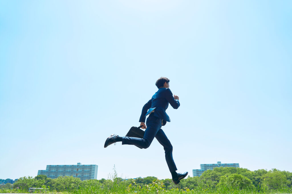 外を走るビジネスマン - SEMICON.TODAY Professional in a blue suit leaps mid-air, holding a briefcase, against a clear blue sky with city buildings in the distance.