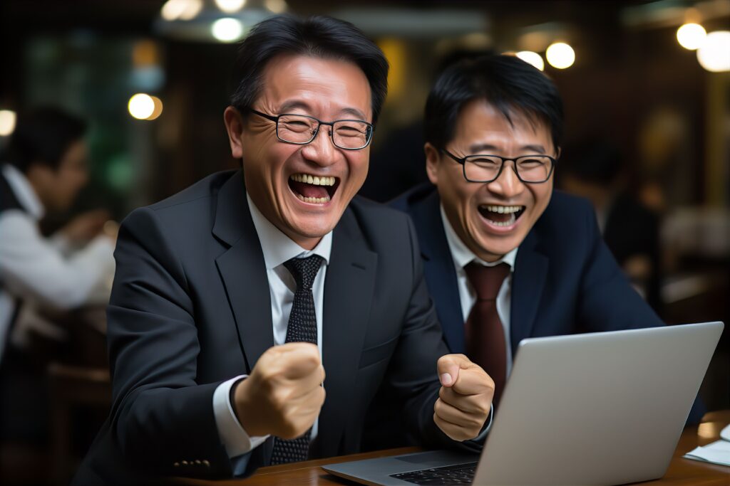 パソコンに向かって喜ぶ日本人の男性ビジネスマンのポートレート写真サラリーマンスーツ若手中堅 - SEMICON.TODAY Two businessmen in suits laugh together at a laptop in a warm, busy office.