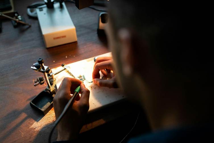 image - SEMICON.TODAY Person soldering a circuit board at a wooden workbench, using a soldering iron and small tools under focused light