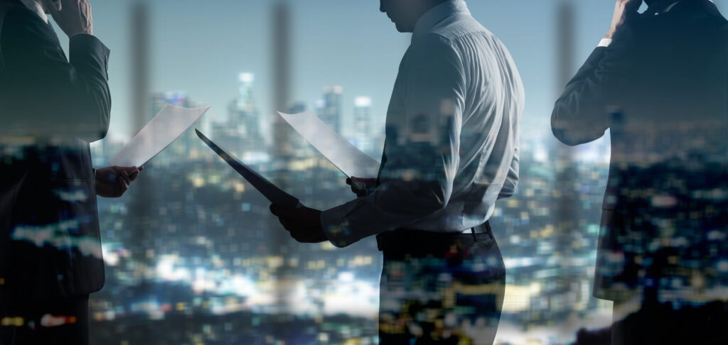 businessman with paper - SEMICON.TODAY Three professionals in suits review documents against a glass wall with a city skyline behind them at night.",