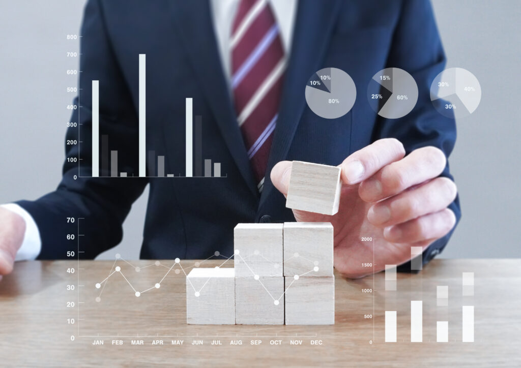 積み木を重ねるビジネスマンとグラフ - SEMICON.TODAY Businessperson in a suit stacks wooden blocks while data charts and graphs float in front, illustrating growth and analysis across months and metrics.