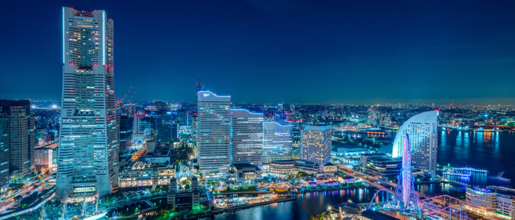 Nighttime city skyline with tall illuminated skyscrapers along a river and neon-blue lights. (Left: tall tower; right: curved waterfront structures)