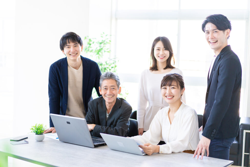 Five professionals in a bright office gathered around a conference table with laptops, smiling at the camera.