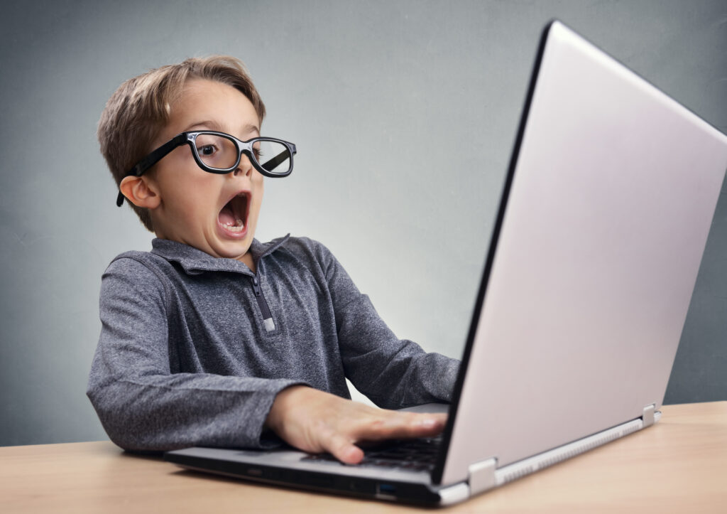 Young boy wearing large black-framed glasses looks astonished while using a laptop on a desk.