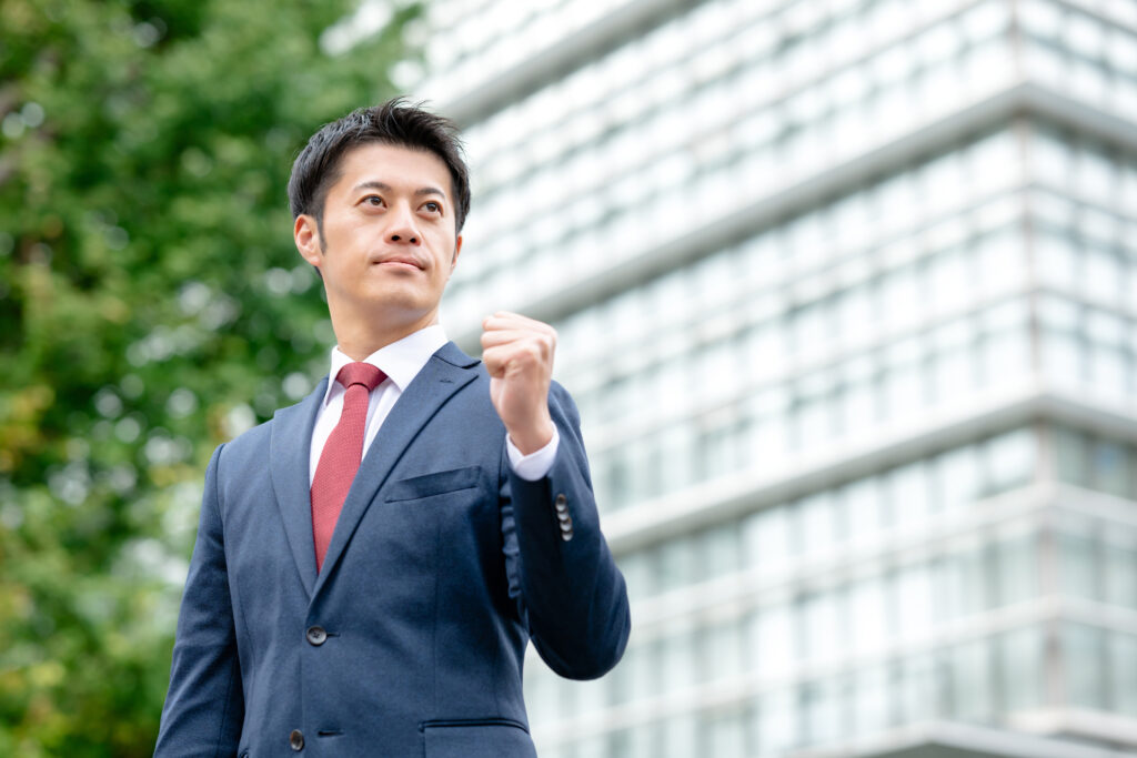 Man in a navy suit and red tie raises his fist outdoors with a glass-building backdrop.