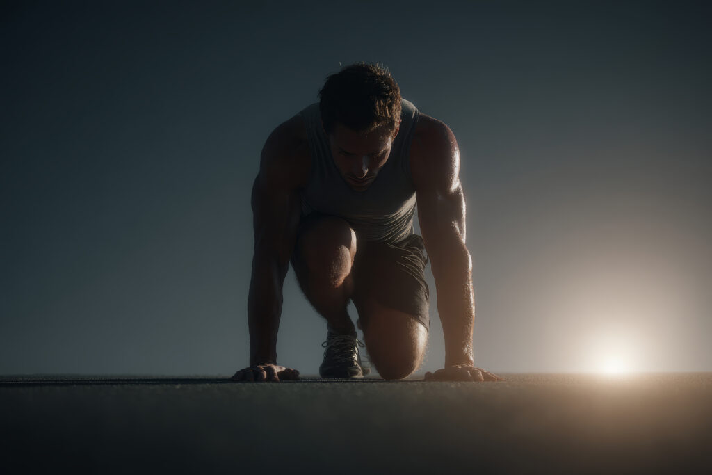 Male athlete in a crouched starting position with hands on the ground, ready to sprint, dusk light behind him on a track surface