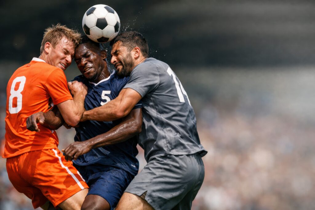 Three soccer players inOrange, blue, and gray shirts struggle for a ball midair during a collision on the field.