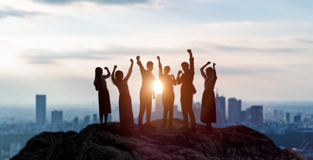 サクセスイメージ - SEMICON.TODAY Silhouetted group of people on a rocky hilltop raising their arms in celebration with a city skyline at sunset behind them.