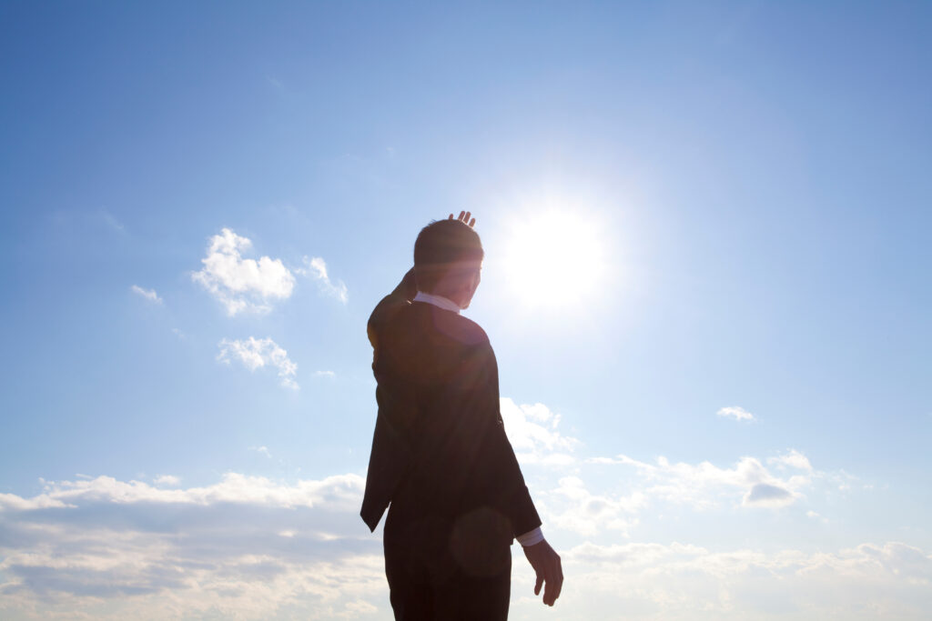 Silhouetted person in a suit raises a hand toward the sun with a bright blue sky behind.