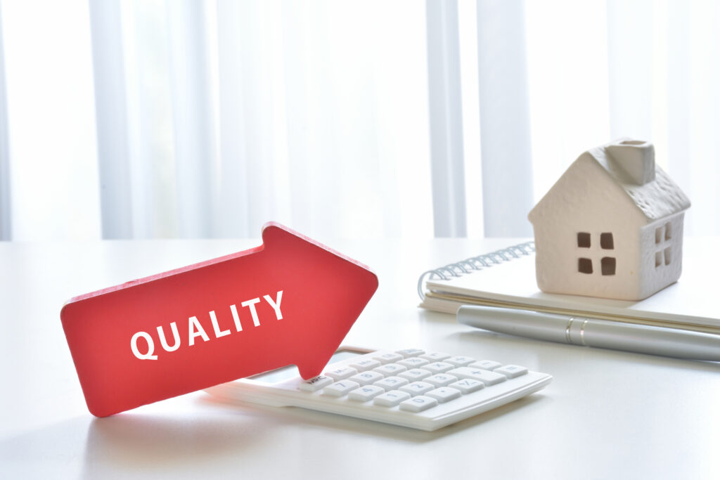 Red sign reading 'QUALITY' on a white desk beside a keyboard, notebook, pen, and a small ceramic house model in the background.