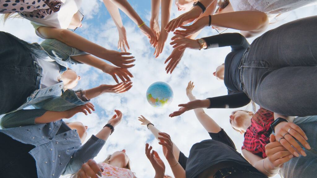 Female student girls standing in a circle toss the world globe up - SEMICON.TODAY Diverse group of people in a circle reaching hands toward a globe at the center under a blue sky, symbolizing teamwork outdoors.