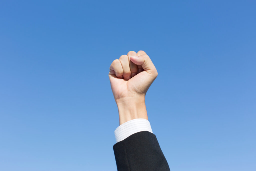 ハンドポーズ - SEMICON.TODAY Raised fist of a person in a dark suit against a clear blue sky.